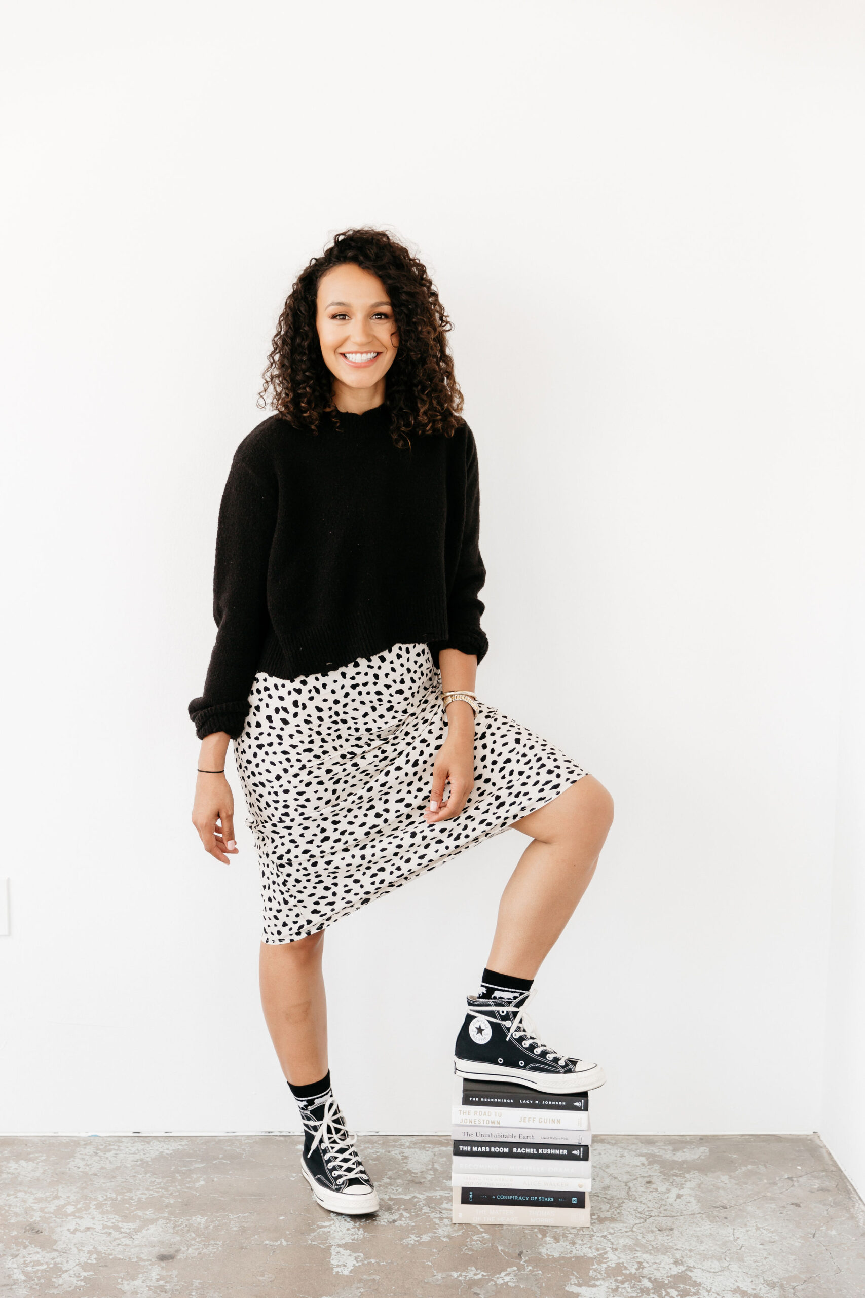 A woman with curly hair, wearing a black sweater, a white skirt with black spots, and black high-top sneakers, stands smiling with one foot on a stack of books against a plain white background.