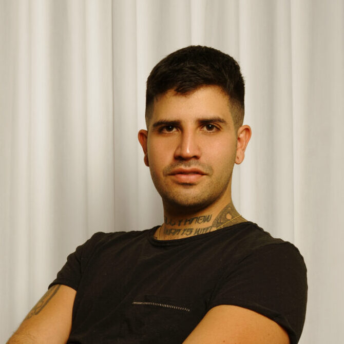 A person with short dark hair sits cross-legged on a textured white chair, wearing a black t-shirt, black pants, and black socks. The background features white curtains and a gray floor.
