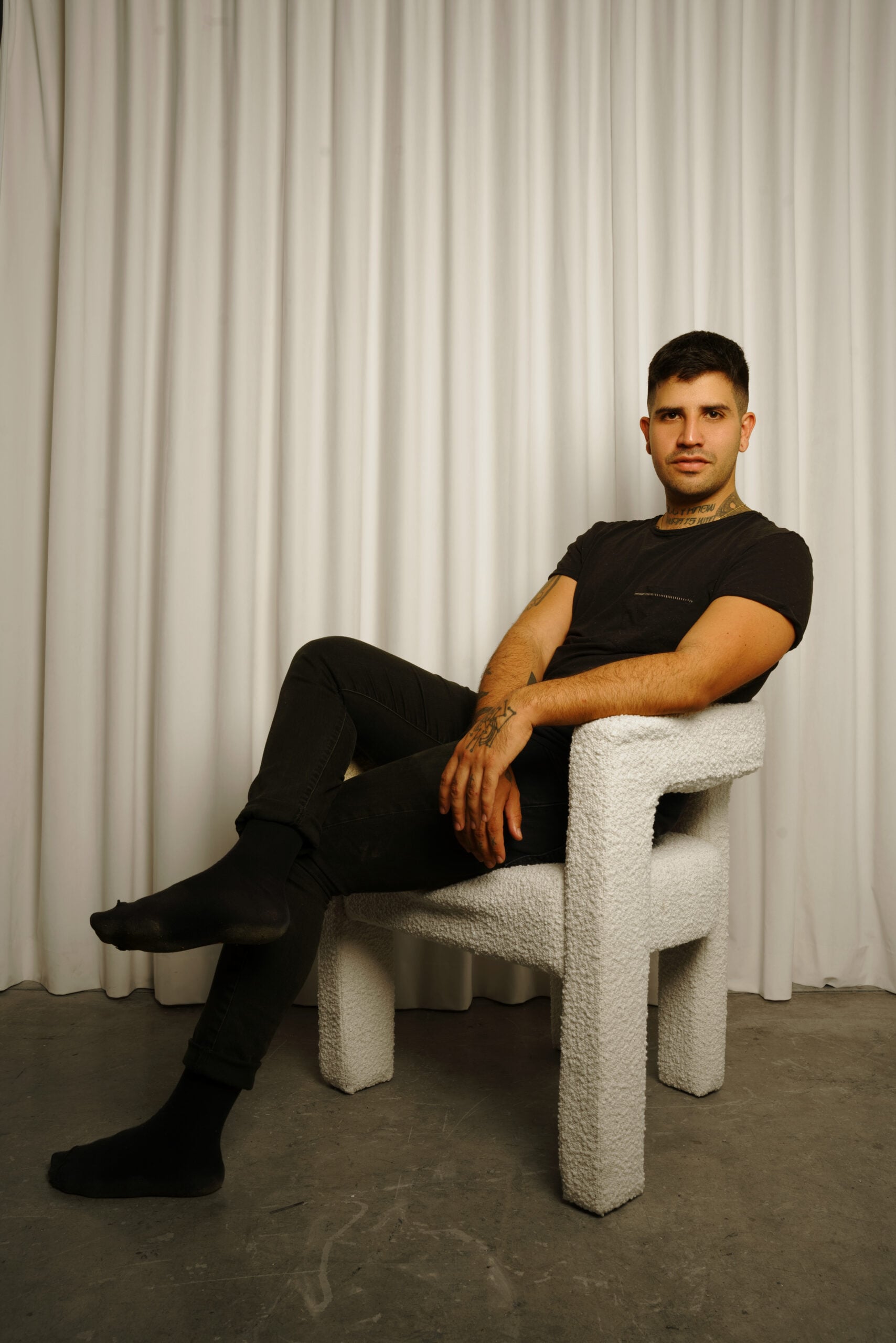 A person with short dark hair sits cross-legged on a textured white chair, wearing a black t-shirt, black pants, and black socks. The background features white curtains and a gray floor.