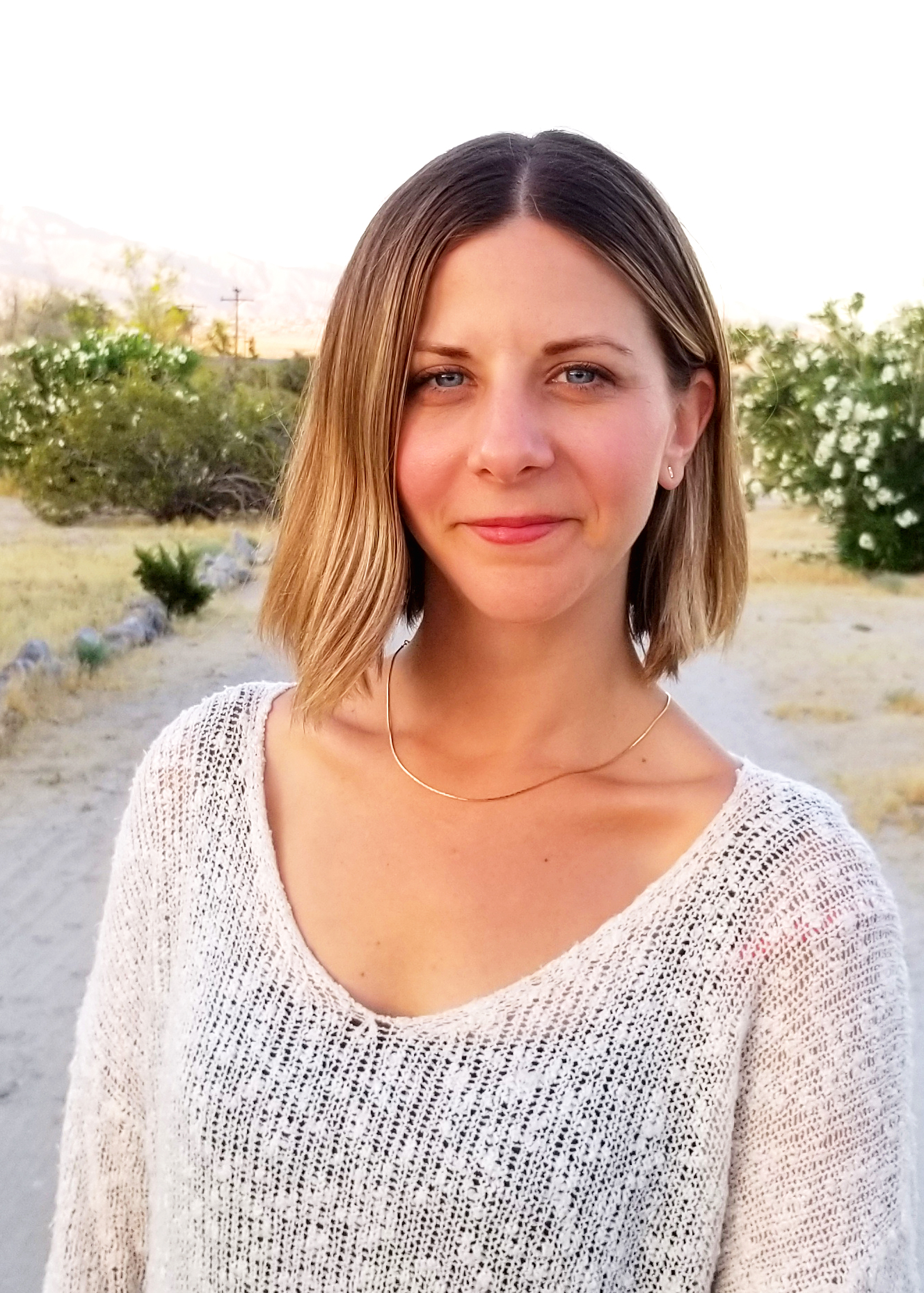 A woman with shoulder-length, straight brown hair stands outdoors on a sunny day, wearing a light-colored knit sweater and a delicate gold necklace. Greenery and trees are visible in the background.