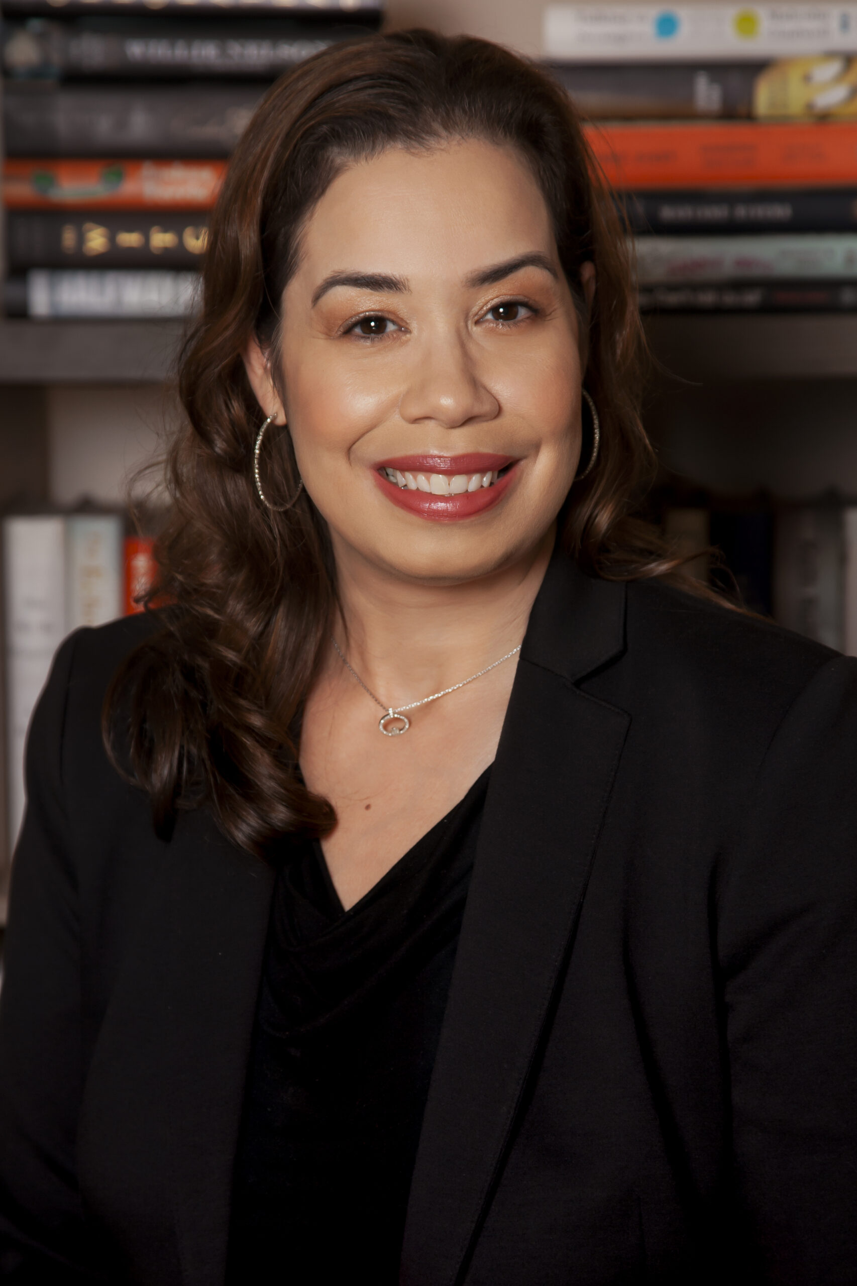 A woman with long brown hair, wearing hoop earrings, a necklace, and a black blazer, smiles at the camera in front of a bookshelf filled with books.