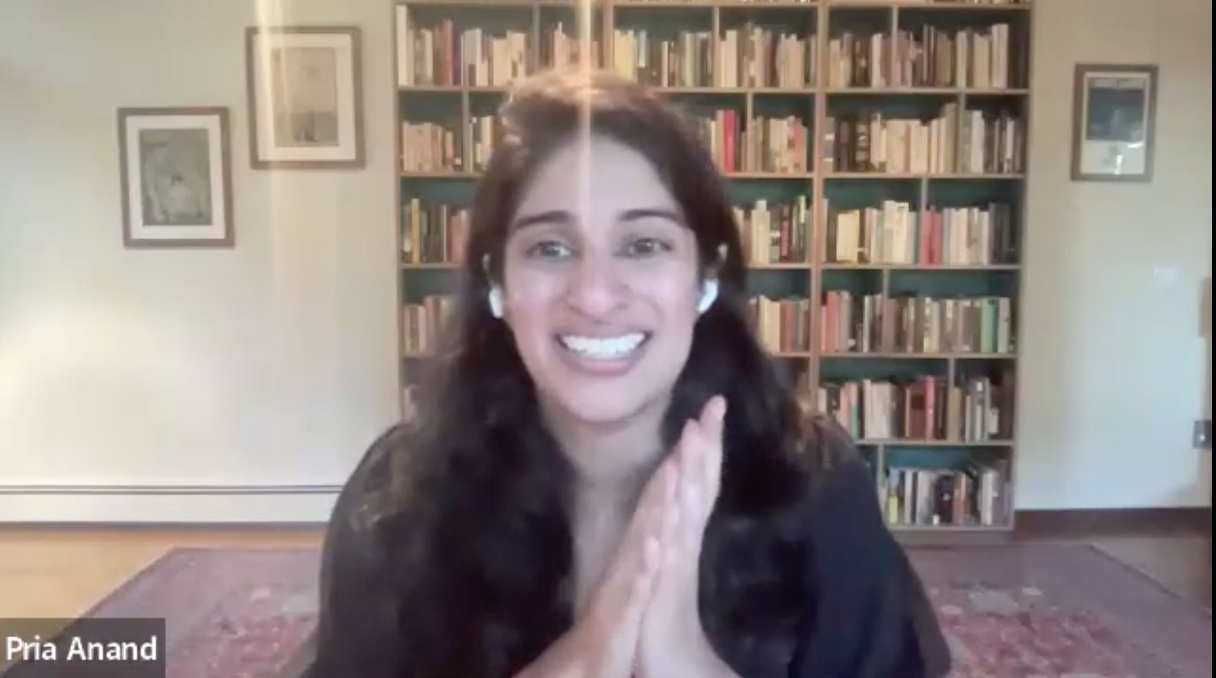 A woman with long dark hair, wearing earbuds and a black top, smiles and clasps her hands together during a video call. She sits in a room with bookshelves filled with books behind her. Her name, Pria Anand, appears on screen.