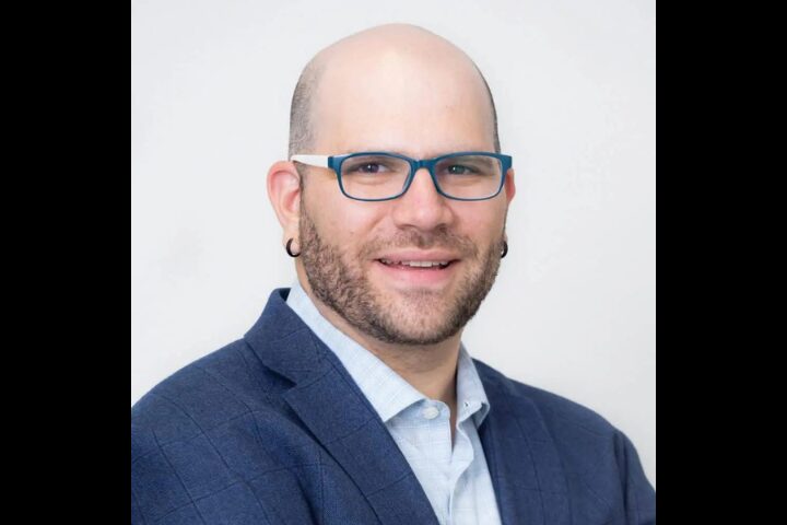 A bald man with a trimmed beard, wearing blue-rimmed glasses, a blue blazer, and a light-colored shirt, smiling slightly in front of a plain light background.