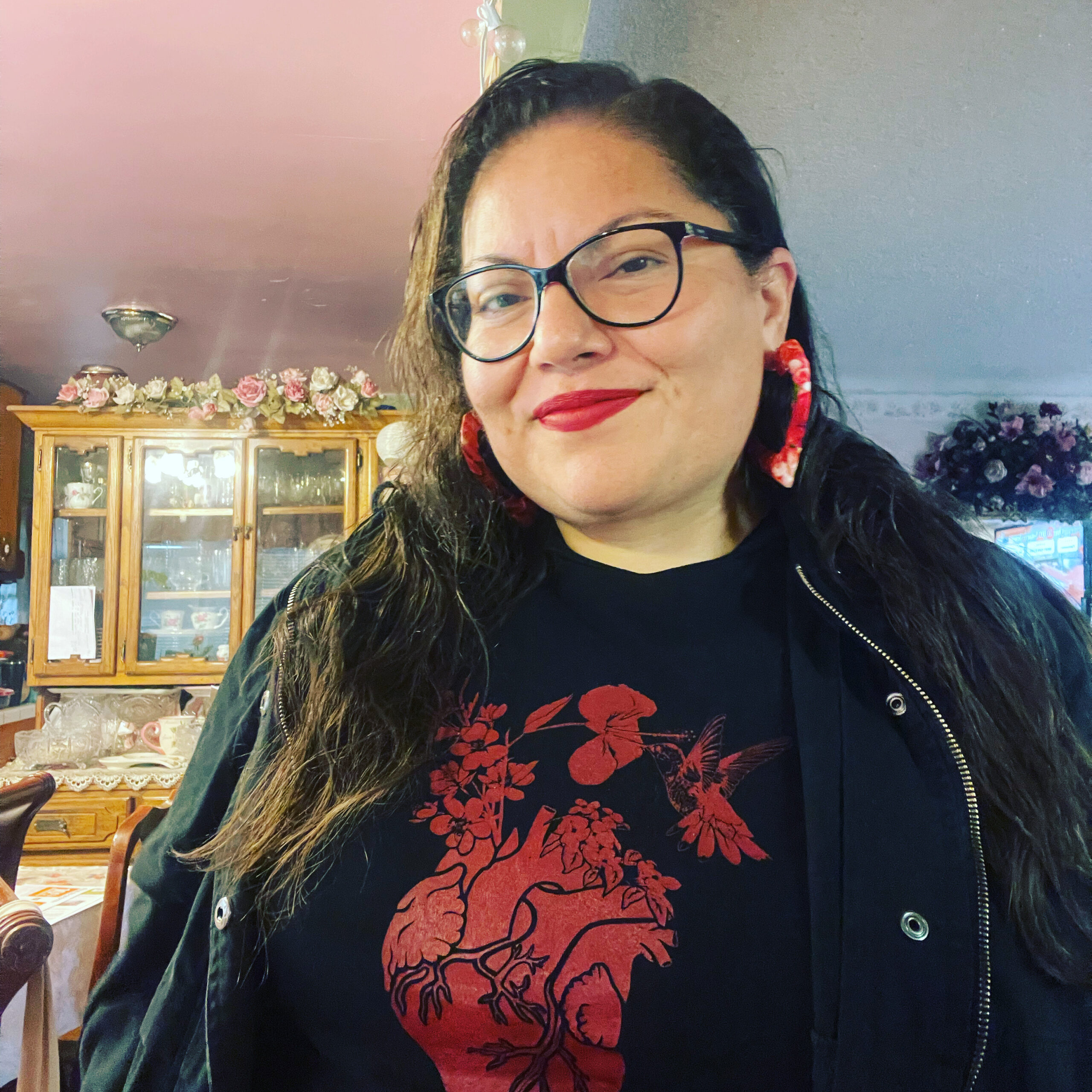 A smiling person with long dark hair, glasses, and red hoop earrings is wearing a black shirt with a red floral and hummingbird design, standing indoors with a wooden cabinet and floral decorations in the background.