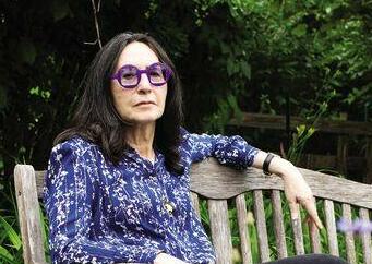 A person with long dark hair and purple glasses sits on a wooden bench outdoors, wearing a blue patterned shirt and black pants, surrounded by green foliage.