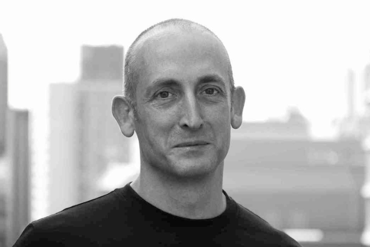 A man with a shaved head wearing a dark shirt stands outside with blurred city buildings in the background. The image is in black and white.