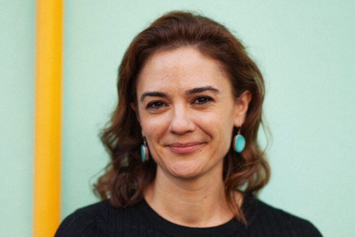 A woman with wavy brown hair and turquoise earrings smiles gently, standing in front of a light green background with a vertical yellow stripe on the left. She is wearing a black top.