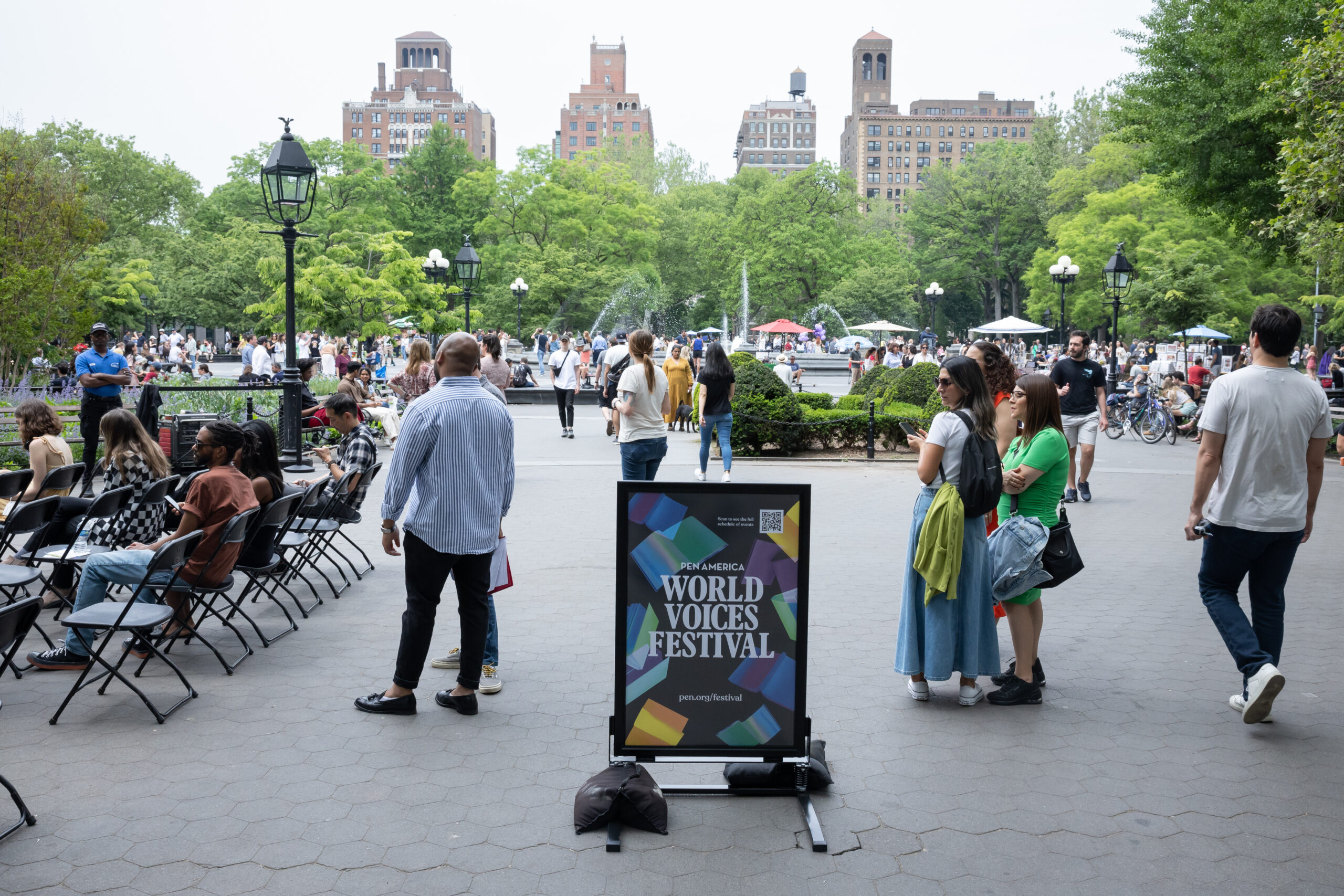 People gather in a park near a sign that reads “World Voices Festival.” Some are seated, others stand or walk. Trees, tall buildings, and a fountain are visible in the background. The atmosphere appears lively and social.