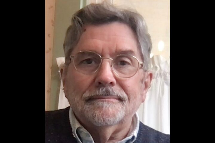 An older man with glasses, gray hair, and a beard is looking at the camera. He is wearing a sweater over a collared shirt. The background appears to be indoors with light-colored walls and curtains.