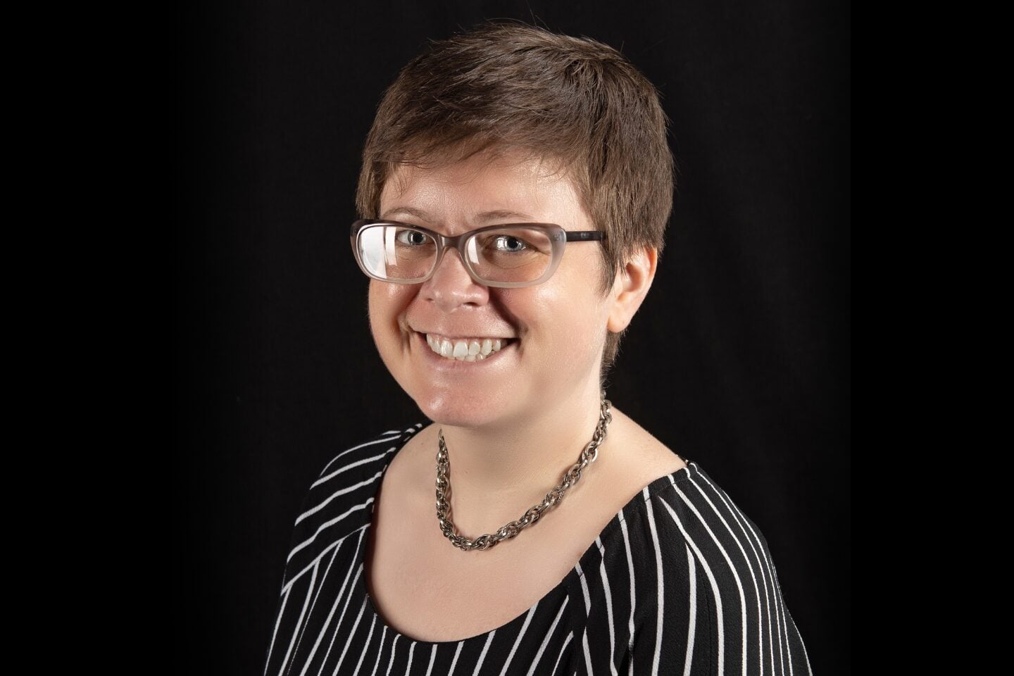 Person with short hair and glasses, wearing a black and white striped shirt and a chain necklace, smiles against a dark background.