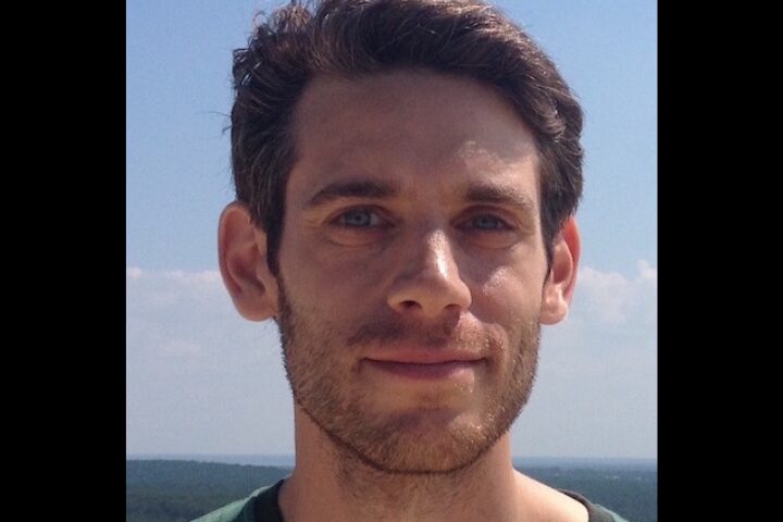 A person with short brown hair and facial hair is facing the camera with a neutral expression. They are outdoors with a clear blue sky and distant horizon in the background.