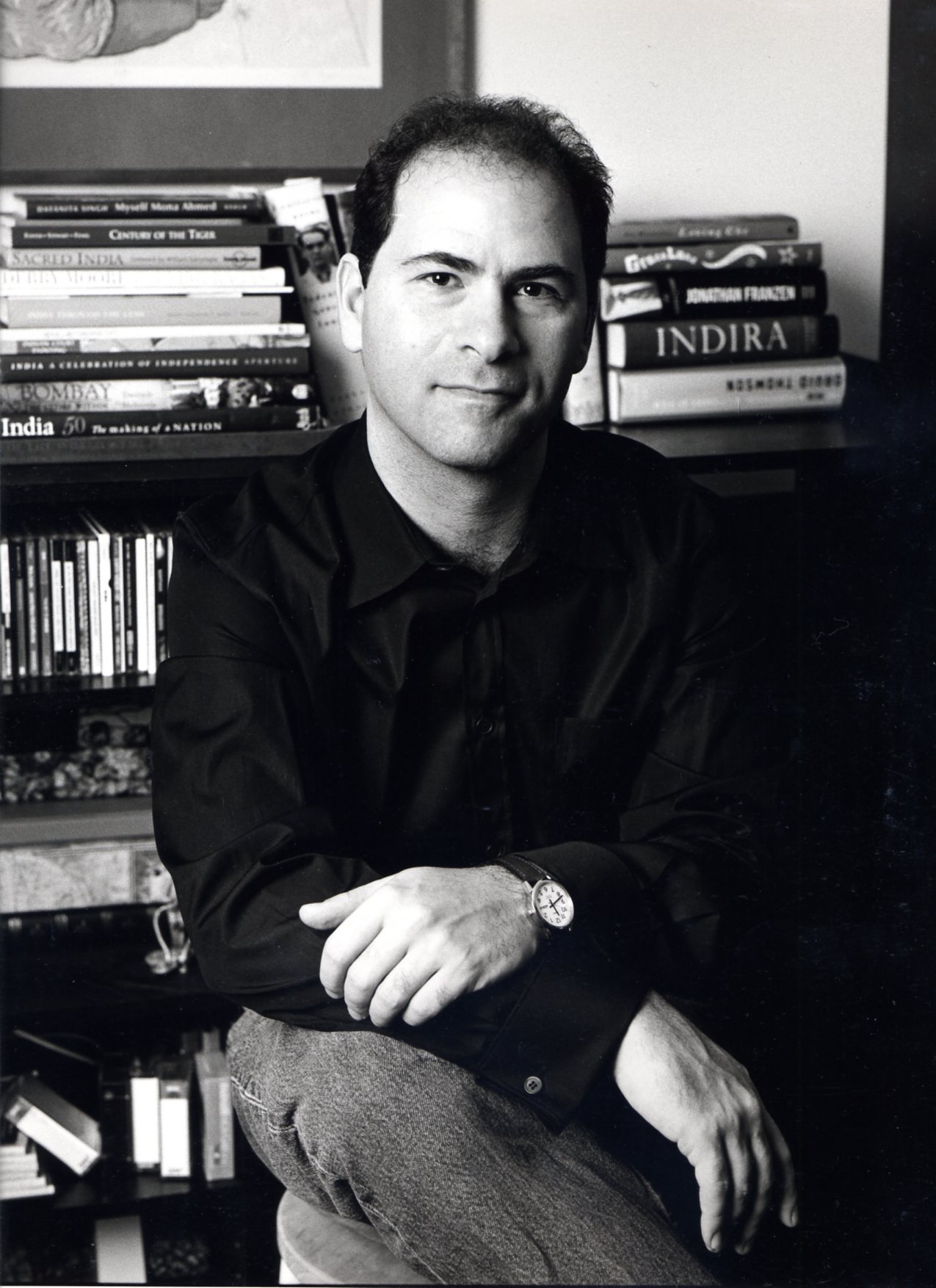 A man in a black shirt sits in front of a bookshelf filled with books. He is resting his arm on his knee and looking at the camera with a neutral expression. The photo is in black and white.