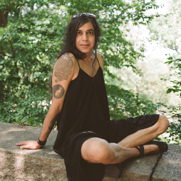 A person with long dark hair sits on a stone ledge in a lush, green outdoor setting. They are wearing a black sleeveless dress, with a tattoo on their upper arm and sunglasses on their head, looking towards the camera with a relaxed expression.