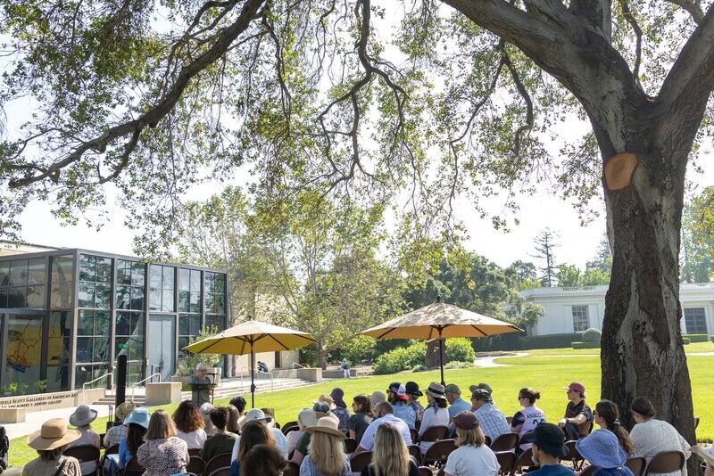A group of people sitting on chairs outdoors under a large tree, facing a speaker near a modern building with glass windows. Sunlight filters through the leaves, and some attendees have umbrellas for shade.