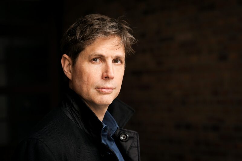 A man with short dark hair, wearing a black jacket over a blue shirt, stands against a dark brick wall. He looks towards the camera, with a serious expression and muted lighting highlighting his face.