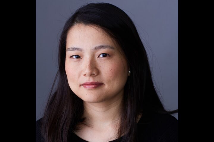 A person with long dark hair and a neutral expression is wearing a black top. The background is a plain gray color.