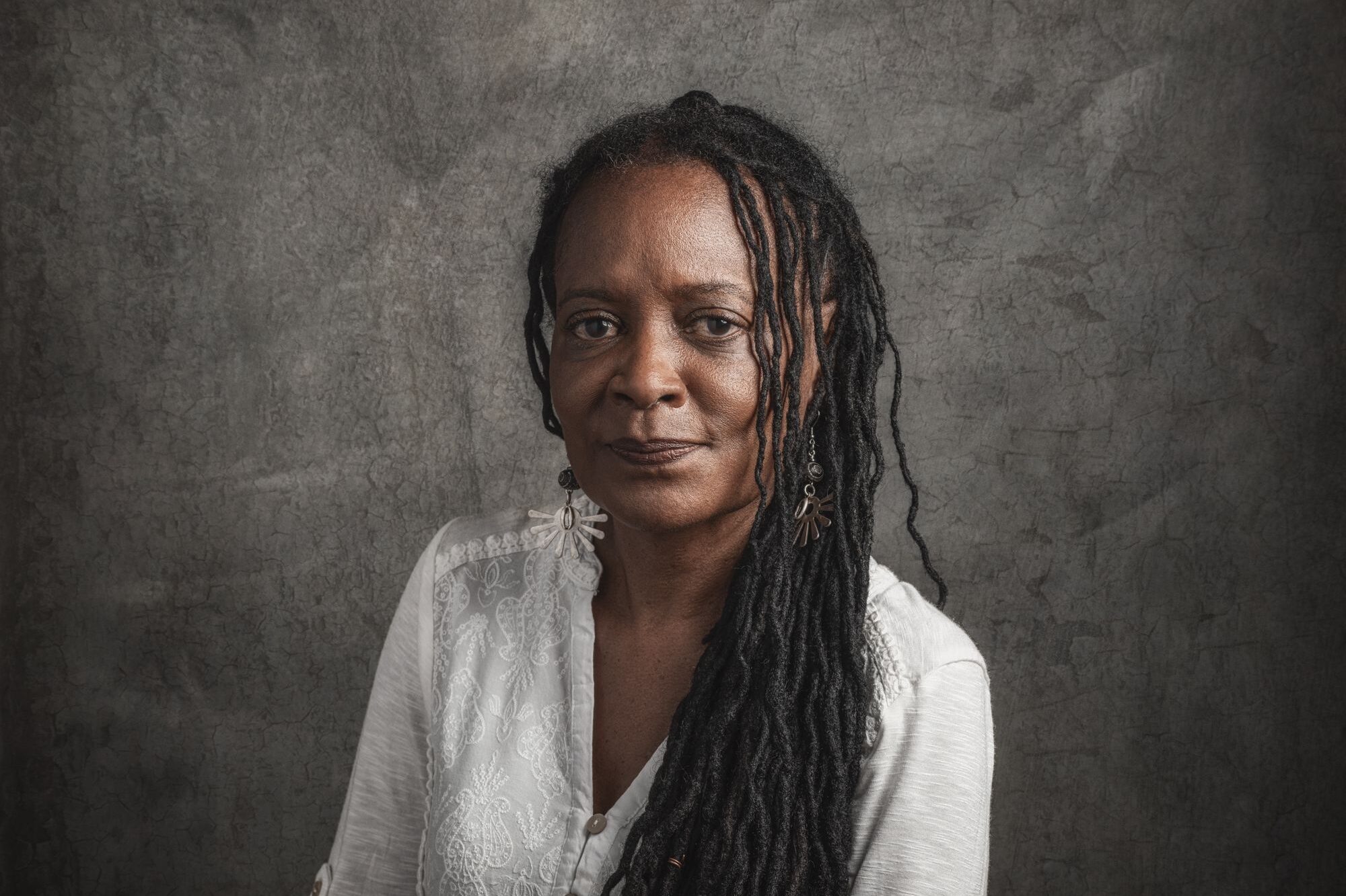 A person with long dreadlocks and a serene expression sits against a textured gray background. They are wearing a white shirt with rolled-up sleeves and are positioned slightly sideways, glancing at the camera.