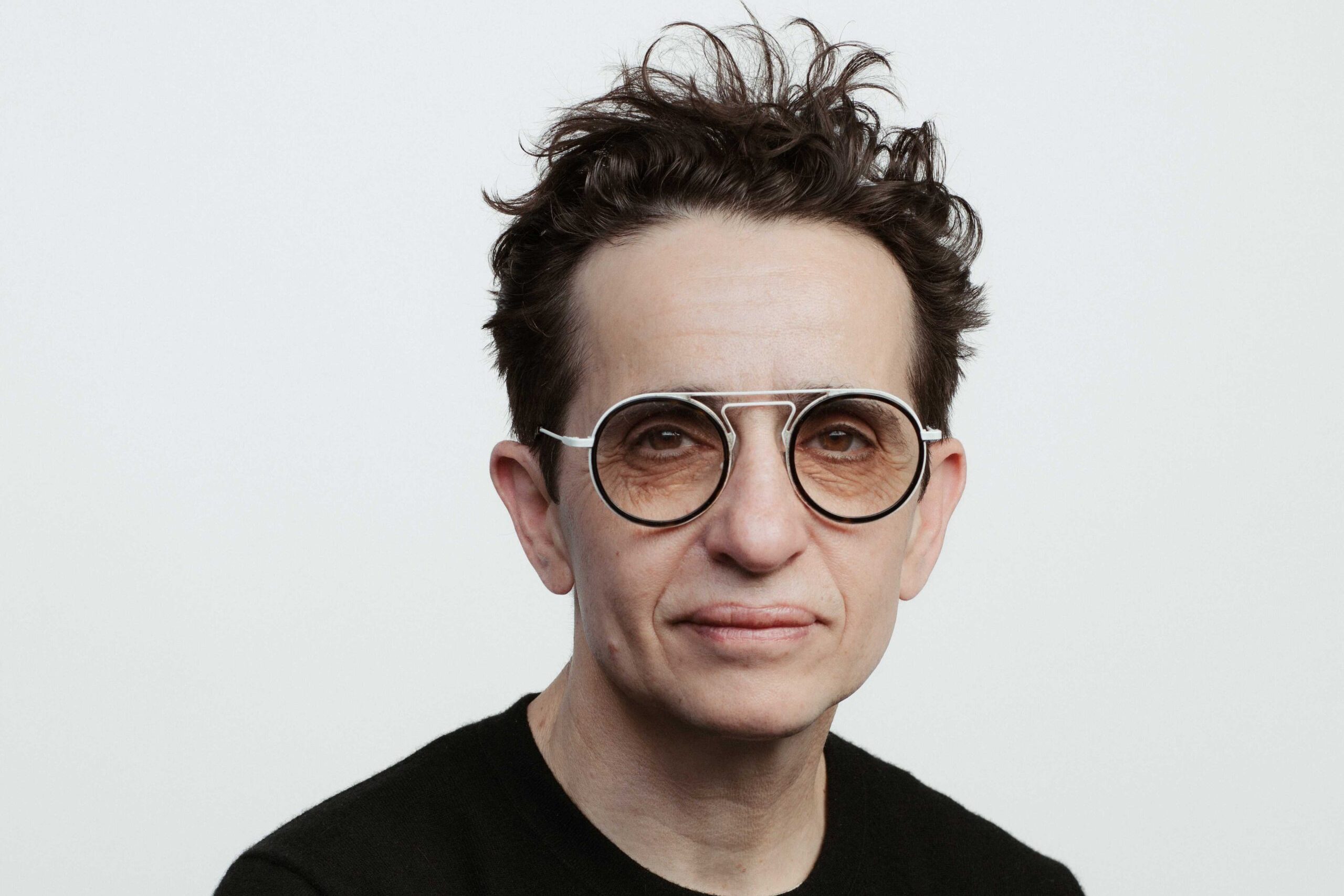 Person with short, curly hair wearing round glasses and a black sweater against a plain background.