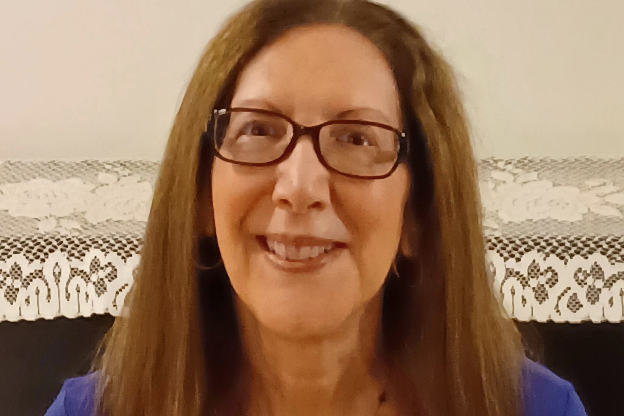 A person with long brown hair and glasses, wearing a blue shirt and a black necklace, is smiling in front of a piano with a lace cover.