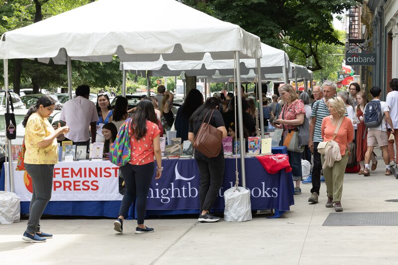 People browsing books at outdoor stalls from Feminist Press and Nightboat Books under white canopies on a city street. Shoppers walk and interact, with trees and a Citibank sign in the background.