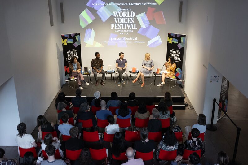 People seated on a stage engage in a panel discussion at the World Voices Festival. The audience is seated below, viewing the event in a room with a large screen displaying festival information.