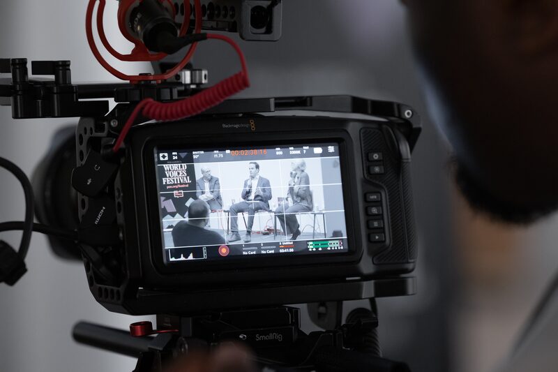 Close-up of a camera screen displaying a panel discussion. Three people are seated on stage with microphones at the World Voices Festival. The camera is framed in the foreground, with a person standing nearby.