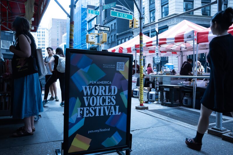 Street scene featuring a PEN America World Voices Festival sign. Red and white tents line the sidewalk with people walking and engaging at stalls. City buildings and street signs for East 12th Street and 4th Avenue are visible.