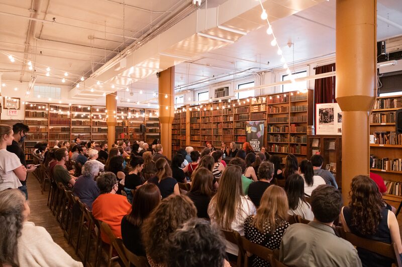A crowded room filled with people seated in chairs, attending an event in a large library. Shelves of books line the walls, and string lights hang from the ceiling, creating a warm atmosphere. A speaker sits at the front, engaging with the audience.