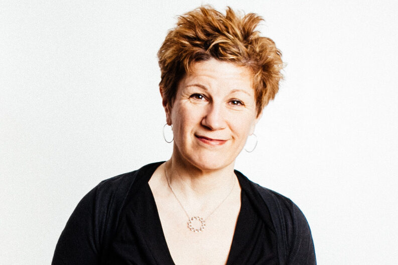 Smiling person with short, tousled hair stands against a white background, wearing a black long-sleeve top and blue jeans. Their hands are behind their back, and they are accessorized with hoop earrings and a necklace.