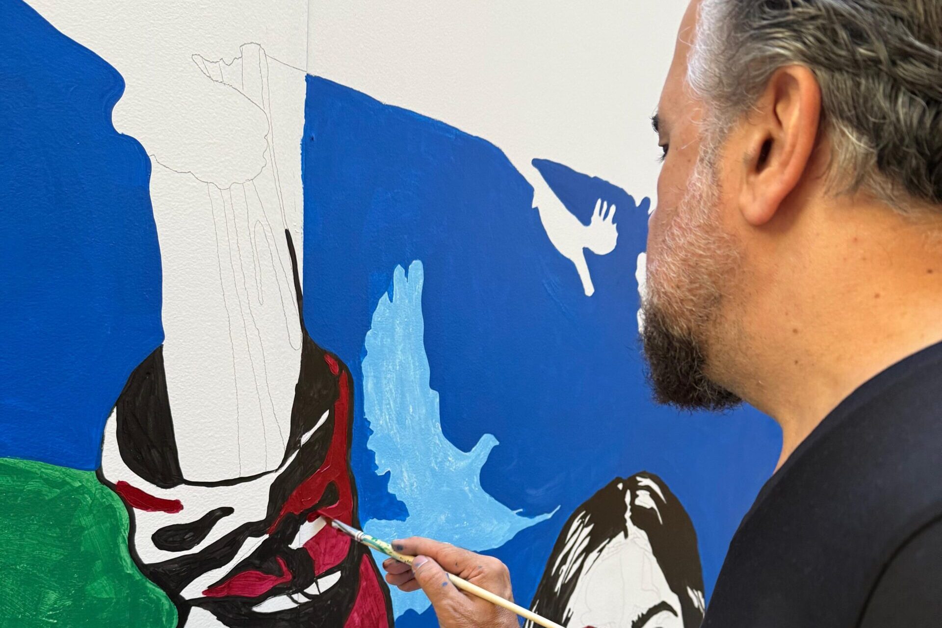 A person is painting a colorful mural featuring an outstretched arm and birds. They hold a paintbrush in one hand and a paint cup in the other. The mural includes vibrant colors like blue, green, and red.