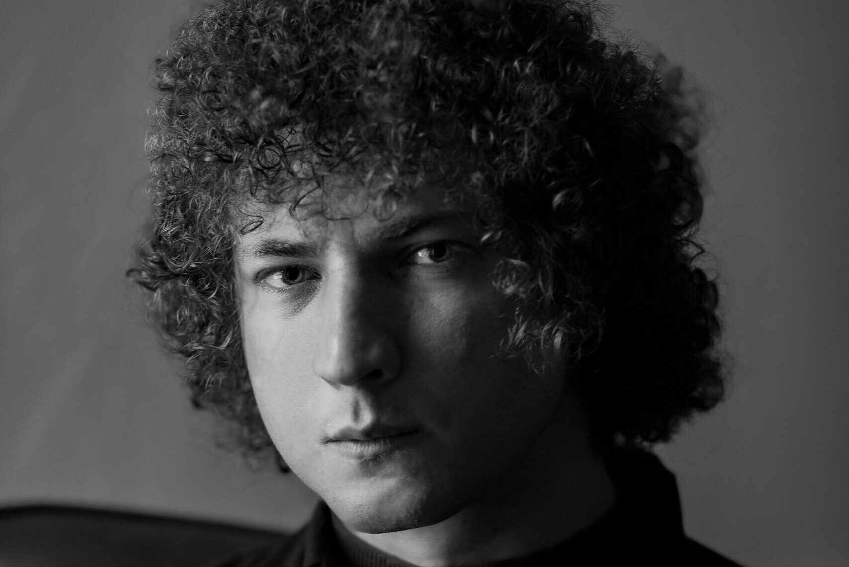 Black and white portrait of a person with curly hair, wearing a dark shirt and turtleneck, sitting against a plain background, looking directly at the camera with a neutral expression.