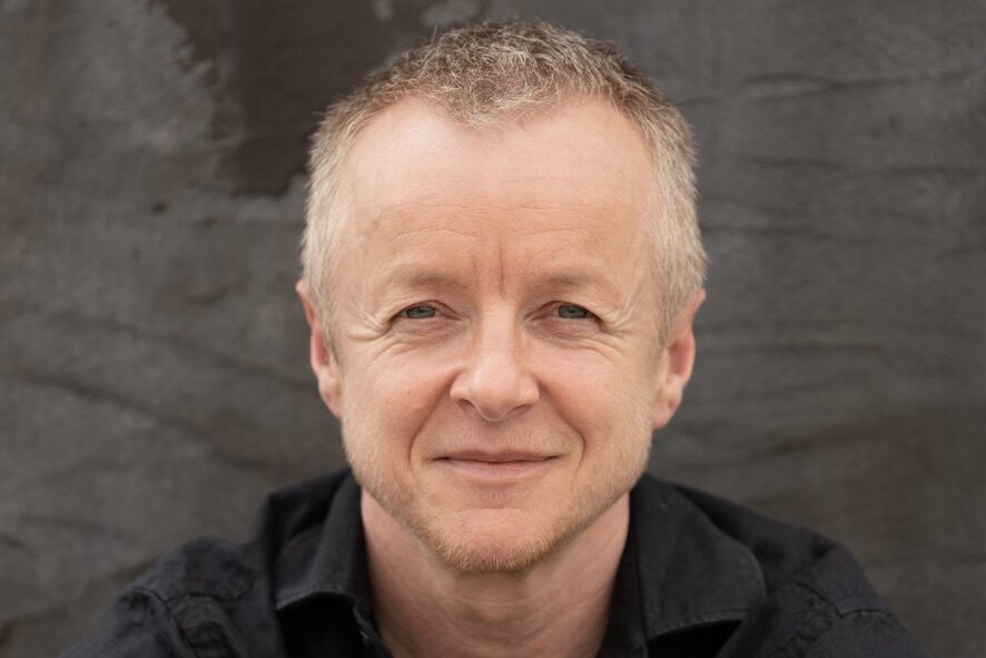 A person with short, light hair and a slight smile wears a black shirt and sits against a textured, dark background.