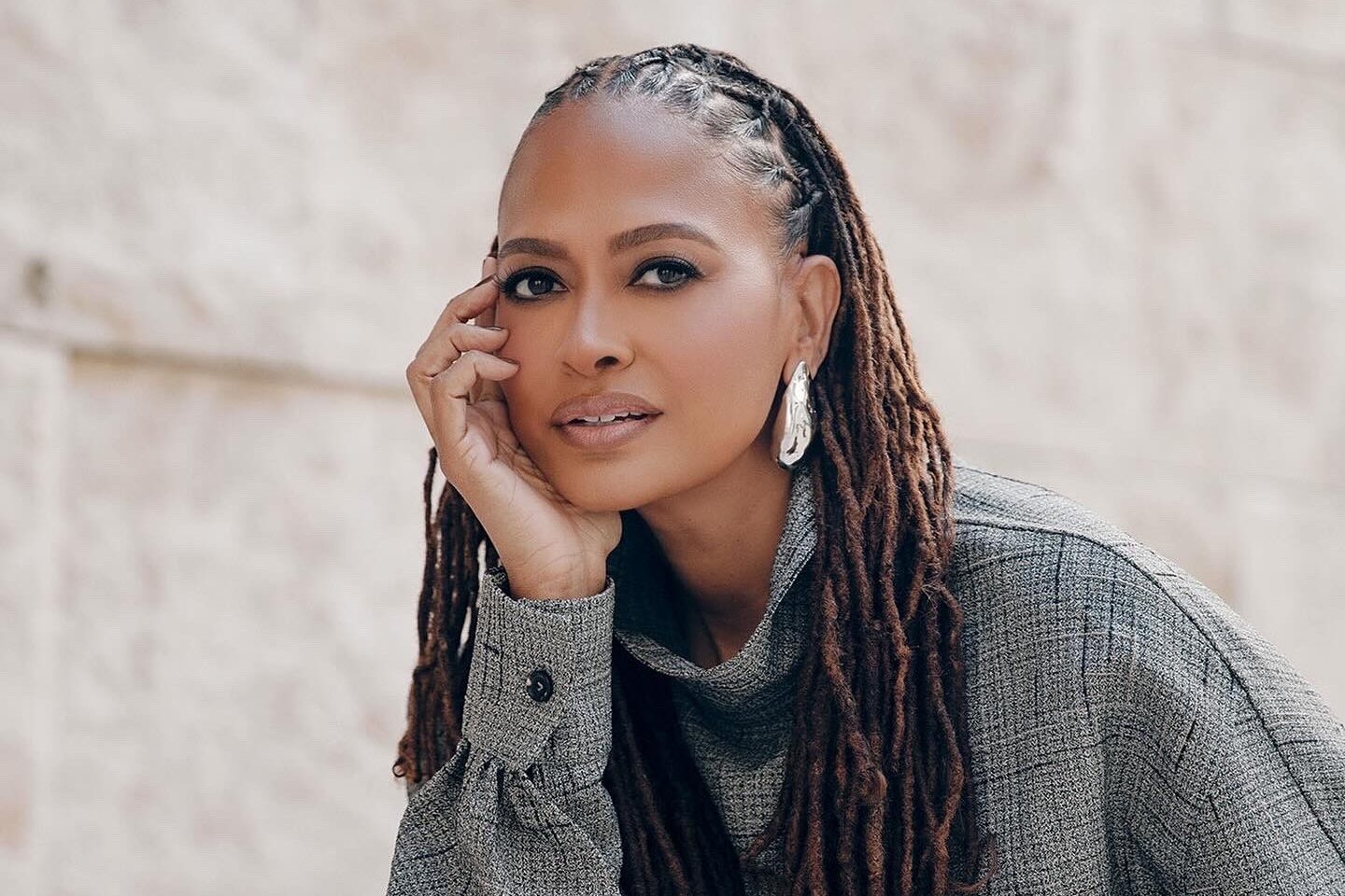 A person with long braided hair sits in a relaxed pose, resting their chin on their hand. They are wearing a gray, textured outfit and large silver earrings, against a light stone wall background.