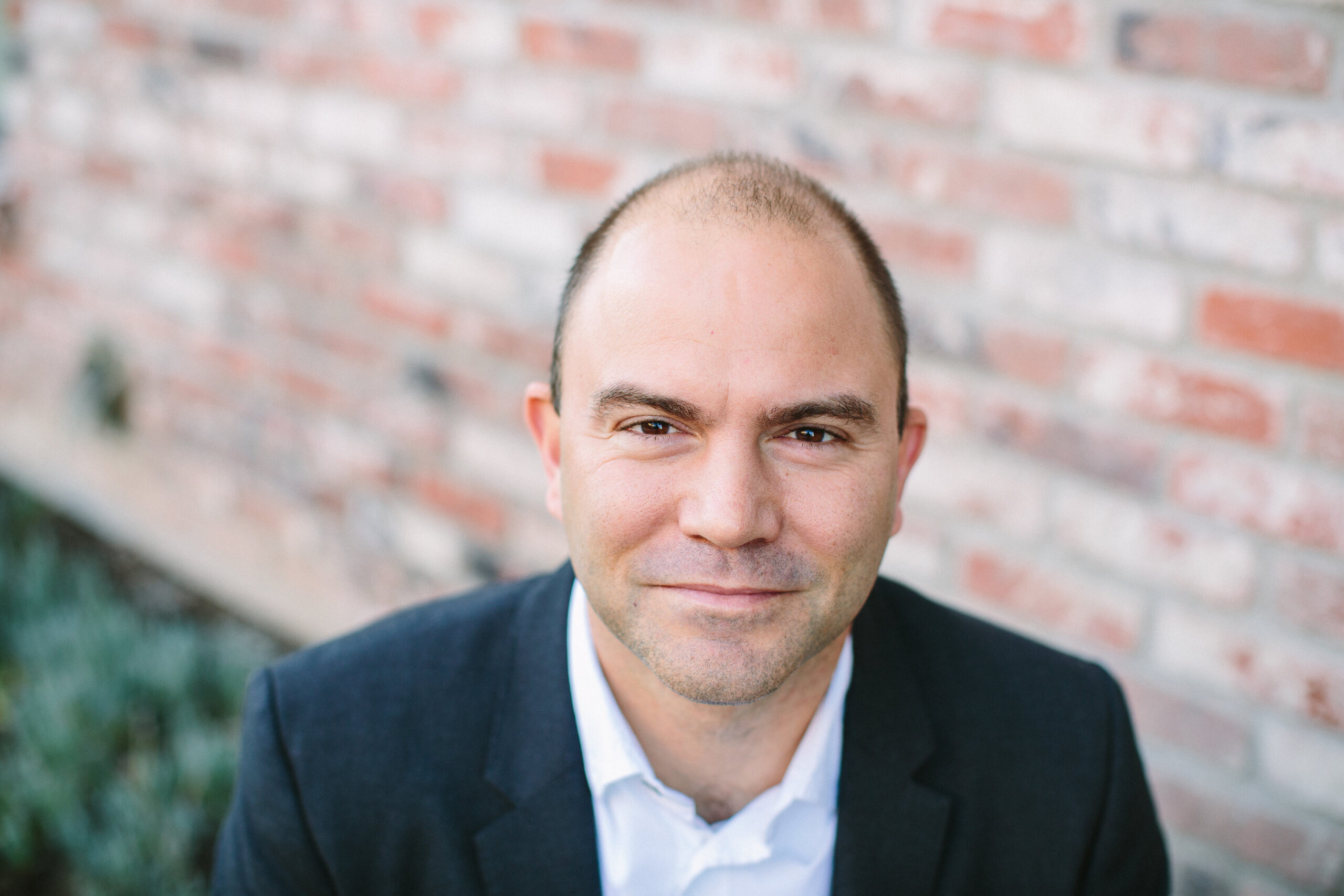 A person with short hair in a black suit and white shirt smiles slightly while looking into the camera. They are standing in front of a blurred brick wall background.