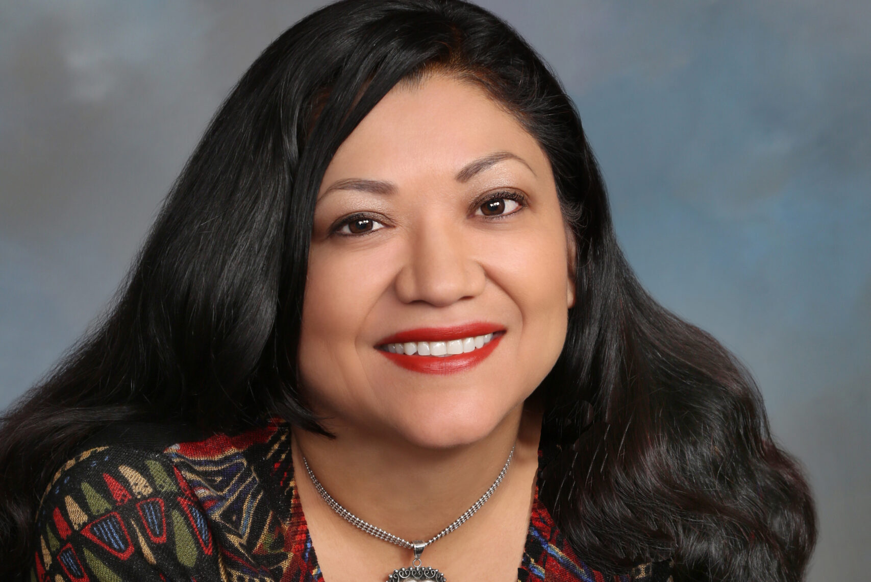 A person with long dark hair and a warm smile is seated with arms crossed, wearing a colorful patterned top and a necklace with a heart-shaped pendant. The background is a soft blend of blue and gray tones.