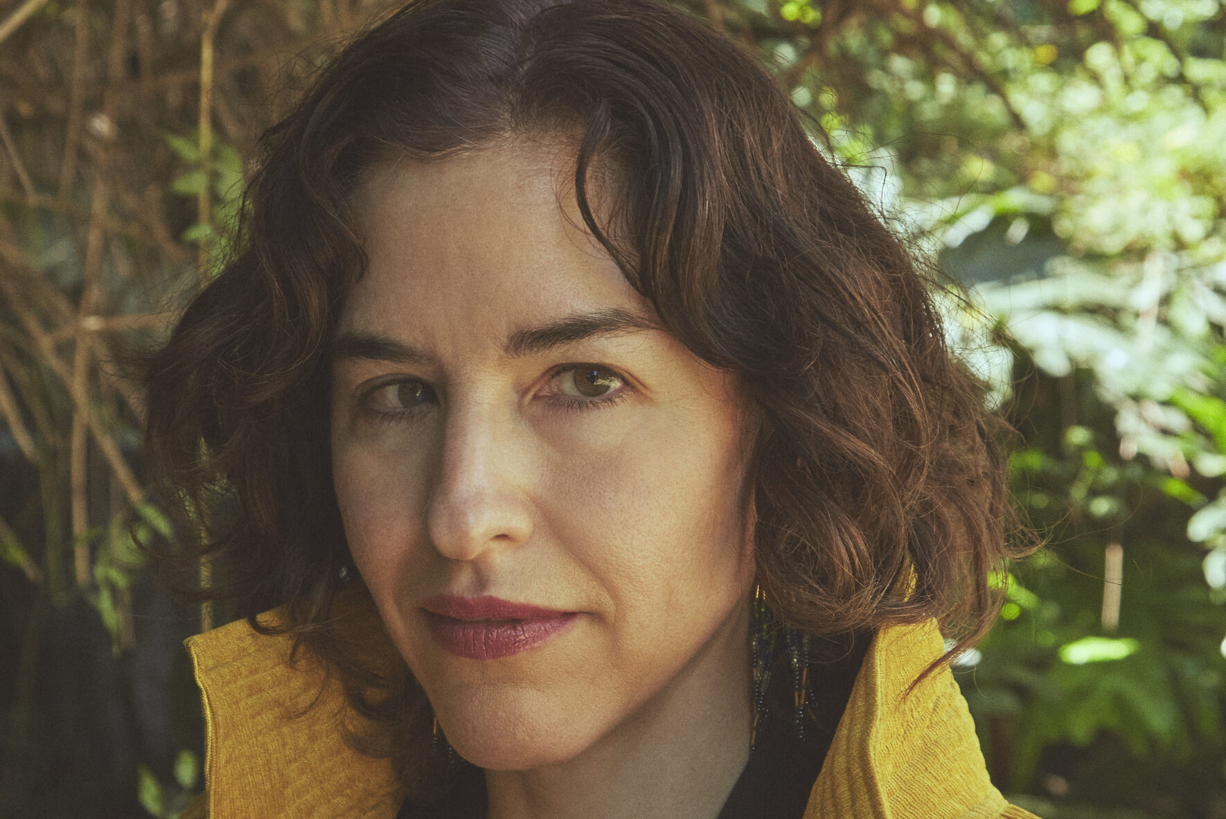 Person with short, wavy dark hair wearing a yellow jacket and black top, standing outdoors with foliage in the background, looking at the camera with a neutral expression.