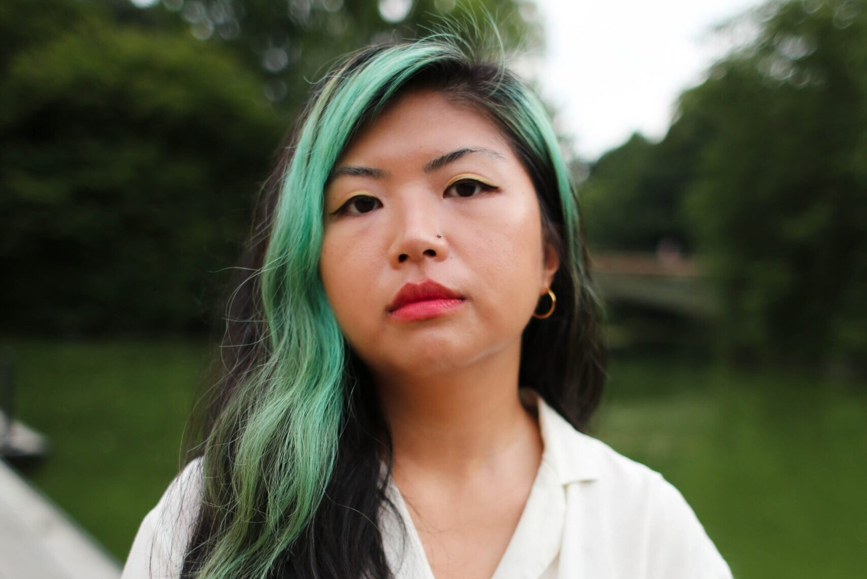 A person with long, black, and green hair stands near a body of water, wearing a white shirt. There are lush green trees in the background. The expression is calm and neutral.