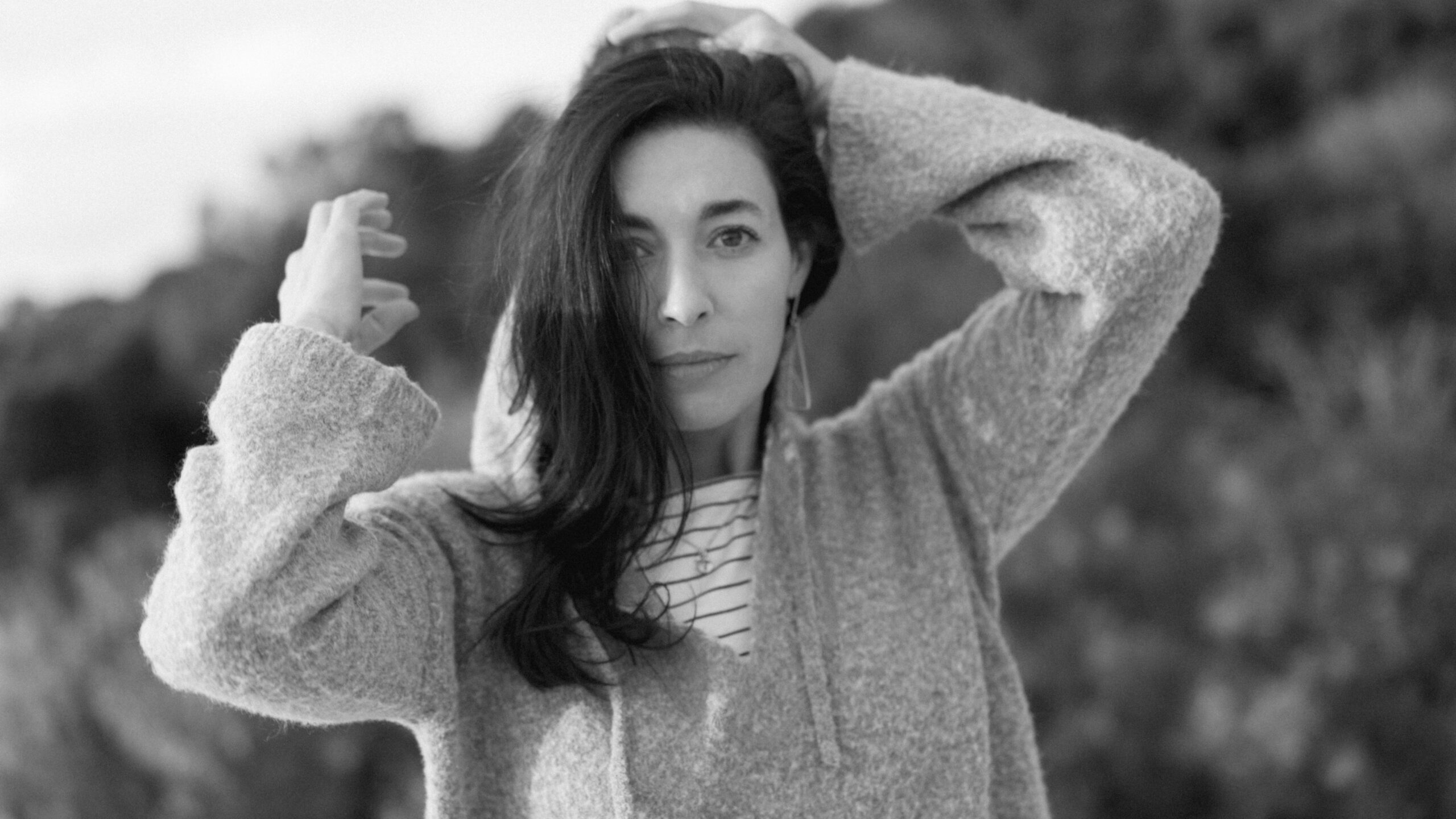 Black and white photo of a person with long hair, wearing a sweater over a striped shirt, looking towards the camera. Their hands are raised, touching their head. The background is blurred, with foliage visible.