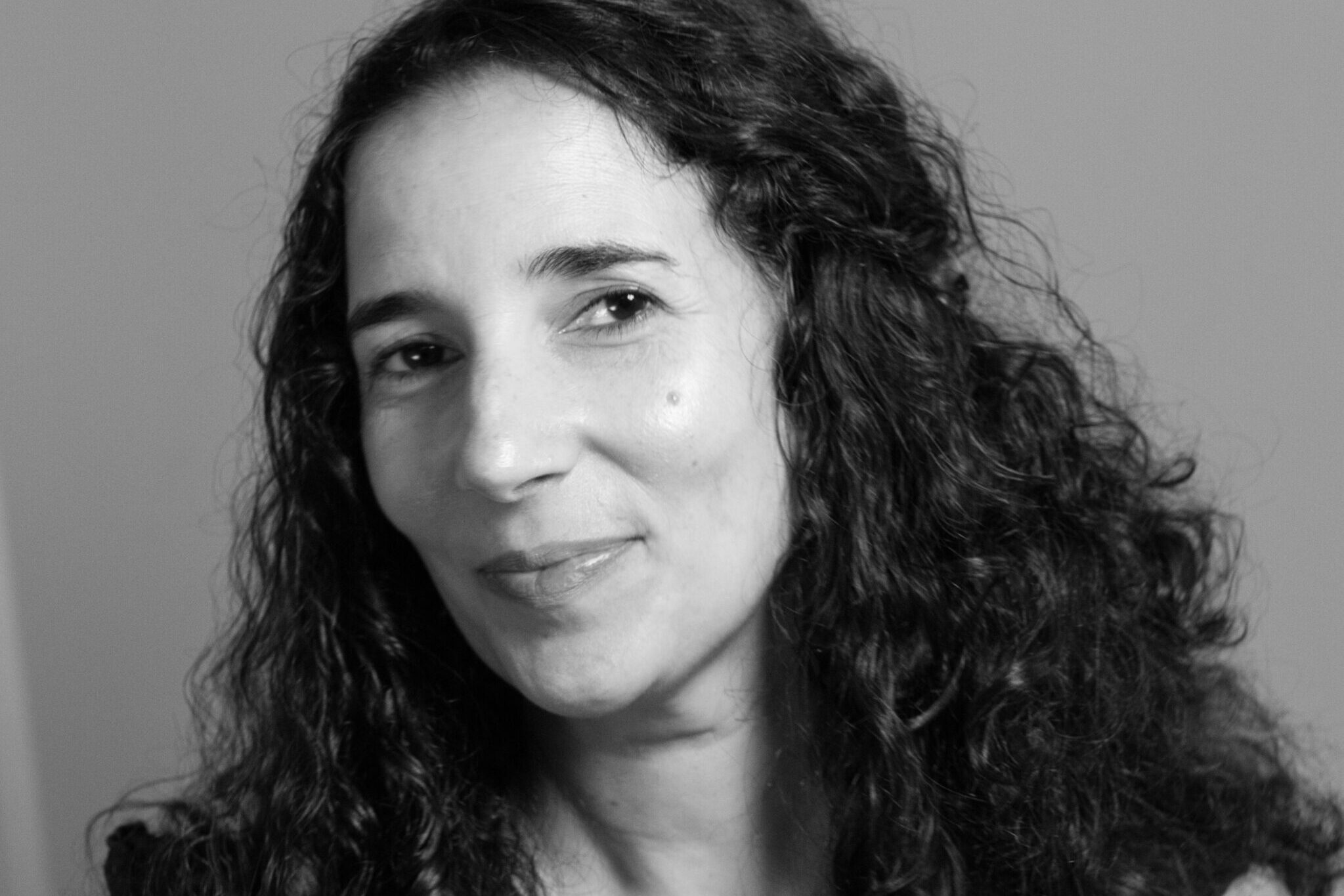 Black and white portrait of a woman with curly hair, smiling slightly. Shes wearing a sleeveless top and looking slightly to the right. The background is plain.