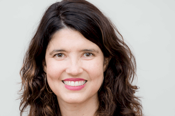 A person with long, wavy brown hair and a bright smile is wearing a black top against a neutral background.