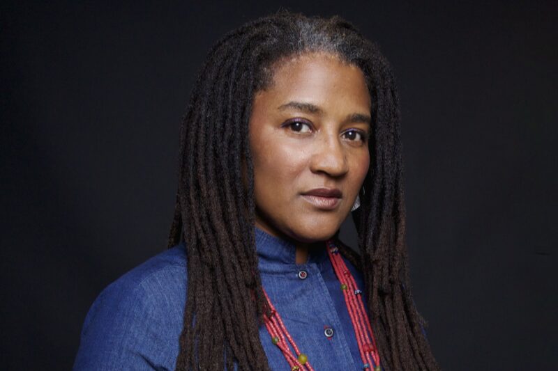 A person with long, dark hair and a hint of gray stands against a black background. They wear a blue top and a red beaded necklace, looking confidently into the camera.