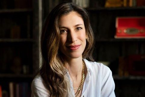 A woman with long, wavy brown hair wearing a white shirt and layered necklaces smiles slightly, seated indoors. The background features books and vintage decor with dim lighting.