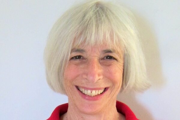A person with short white hair is smiling warmly. They are wearing a red collared shirt and a necklace. The background is plain and light-colored.