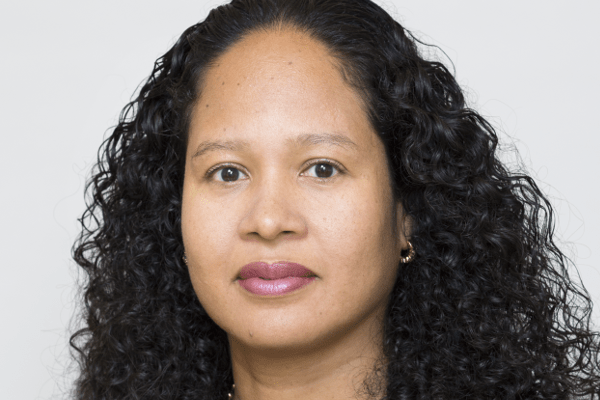 A woman with long, curly black hair is wearing a black top and a gold necklace. She is looking directly at the camera against a plain, light gray background.