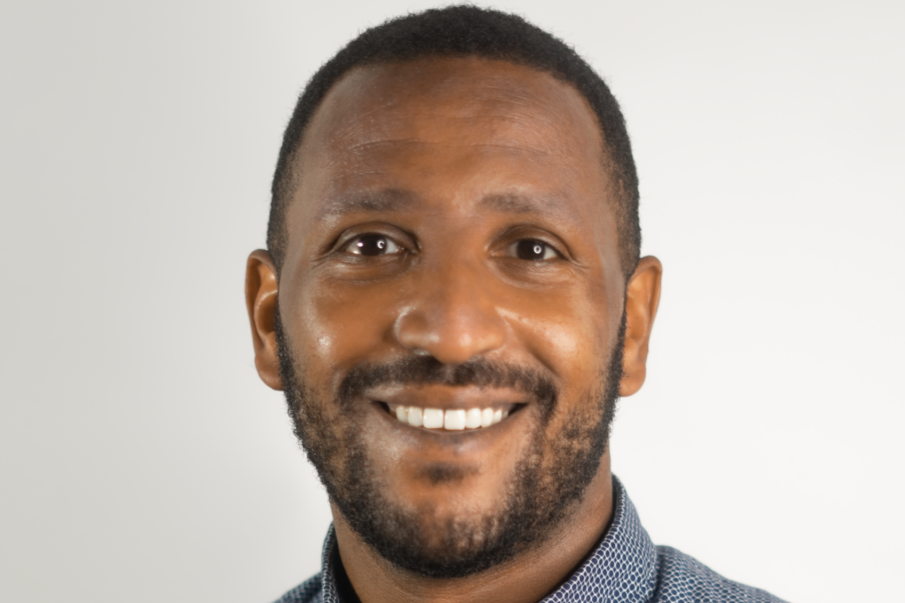 Smiling person with short hair and a beard, wearing a blue and white patterned button-up shirt, standing against a plain white background.