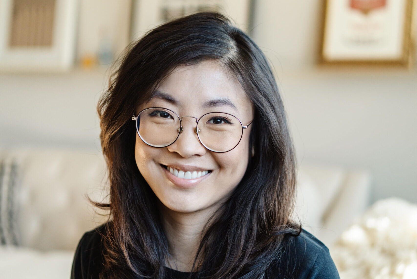 A smiling person with long dark hair and glasses is wearing a black top. They are sitting in a room with framed pictures on a light-colored wall in the background.