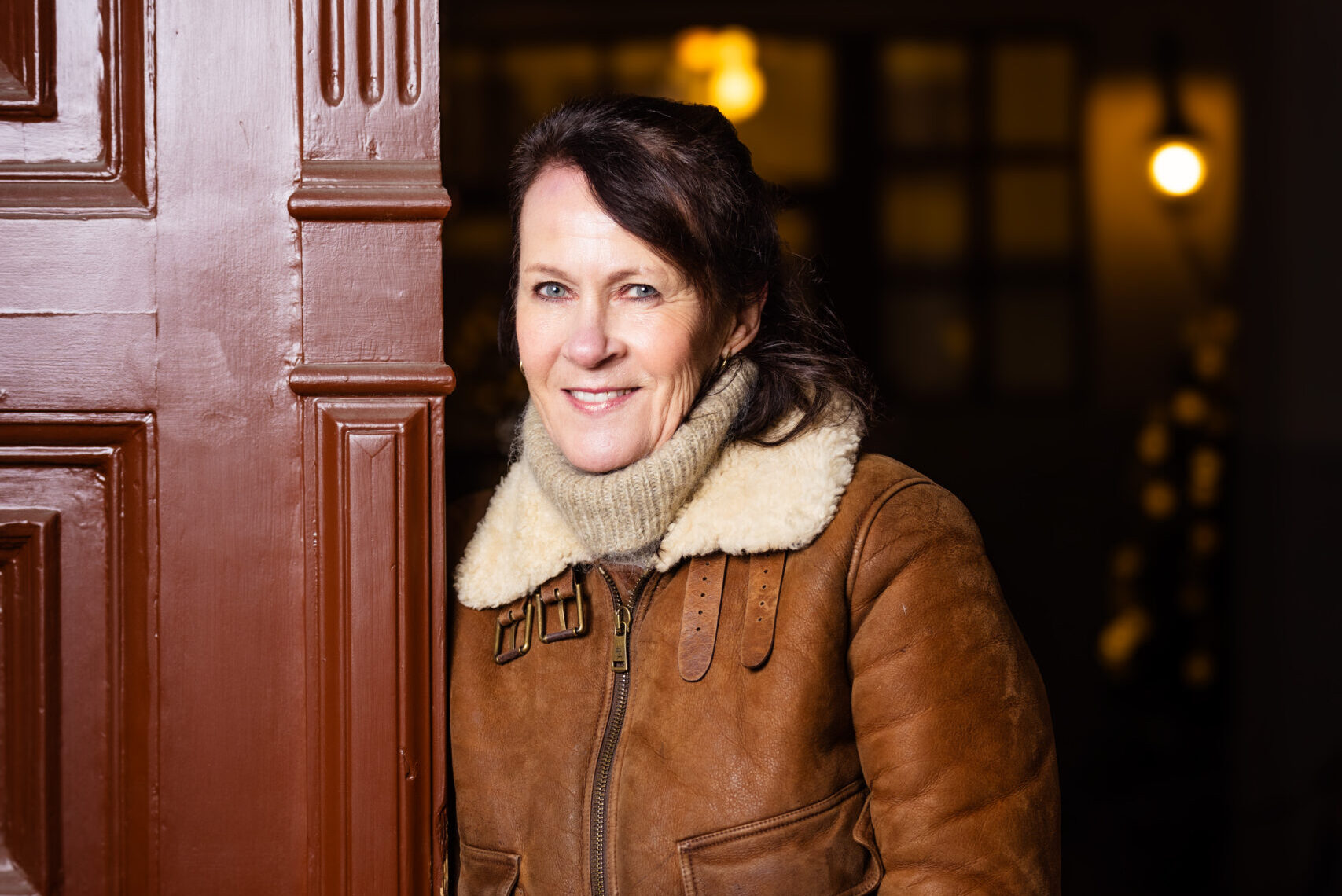 A person stands in a doorway with a wooden door ajar. Theyre wearing a brown, shearling-lined jacket, gloves, and a turtleneck sweater. The background is dimly lit with soft, warm lighting.