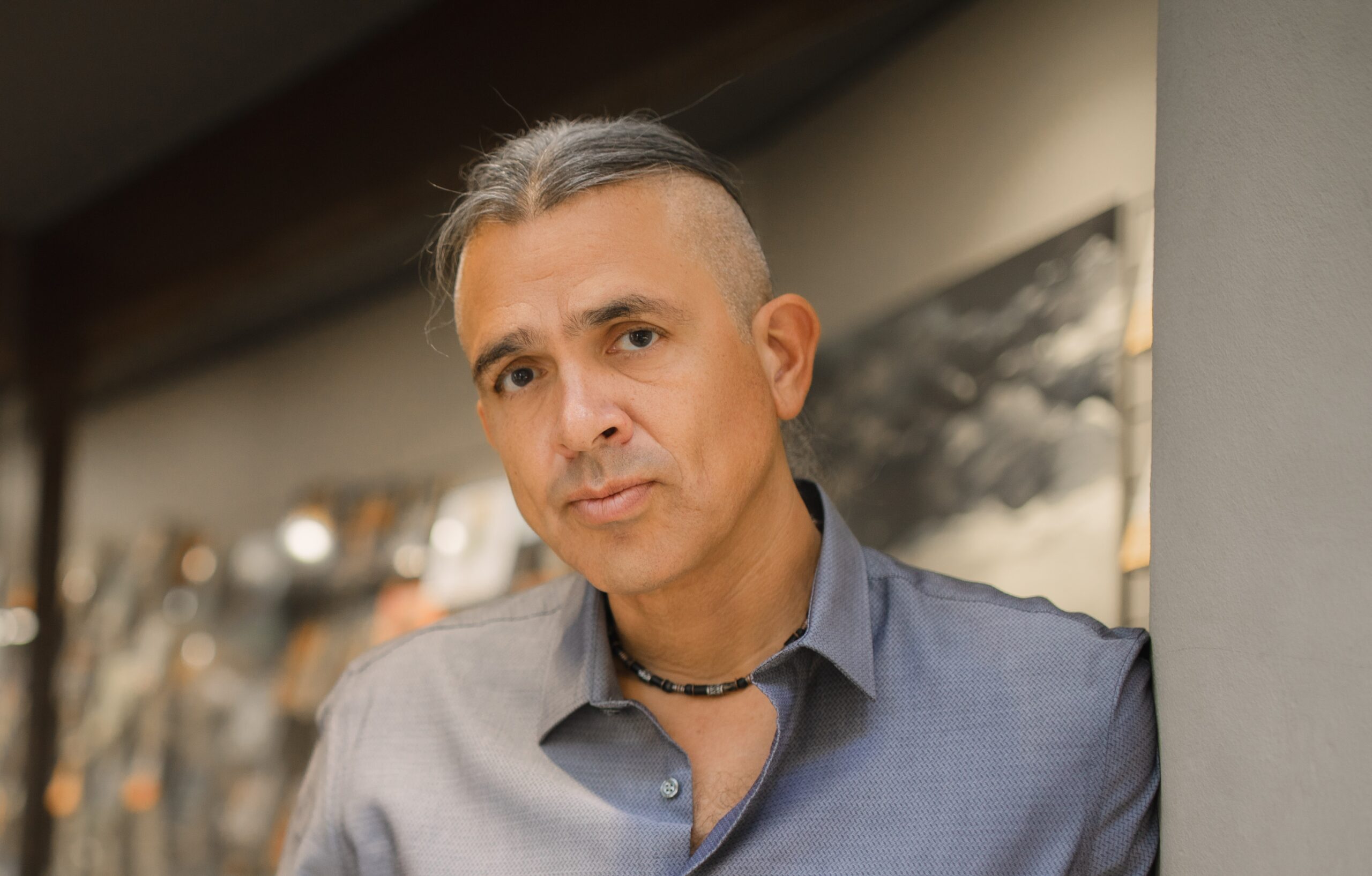 A person with silver hair styled in a top knot and shaved sides leans against a wall. They are wearing a blue button-up shirt and a necklace, gazing directly at the camera with a neutral expression. The background is softly blurred.