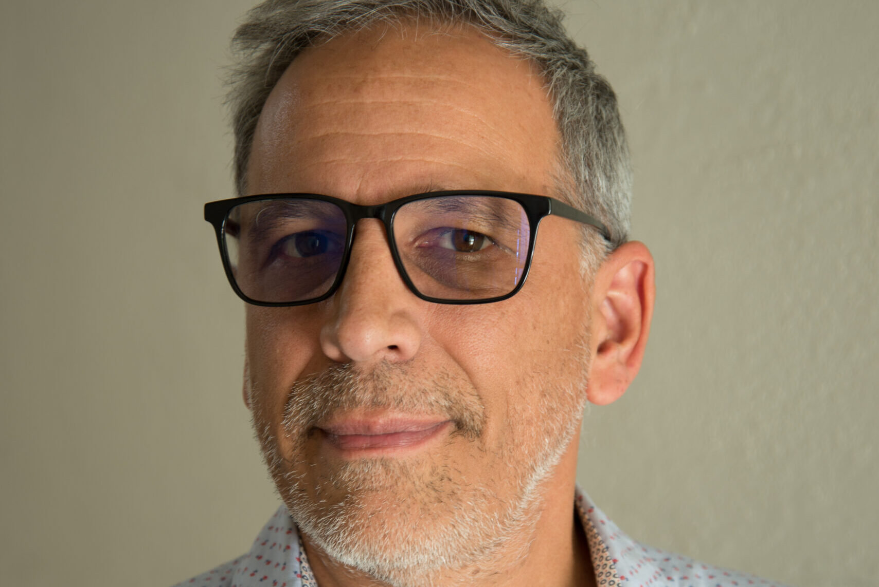 Man with short gray hair and glasses, wearing a light blue shirt with small heart patterns and a white undershirt, stands against a plain light background.