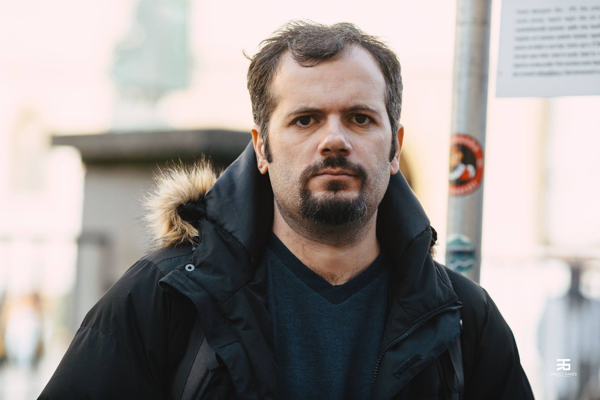 A man with short dark hair, a goatee, and a dark jacket with a fur-lined hood stands outdoors. A pole with stickers and a blurred background of a building are visible. The mood is neutral.